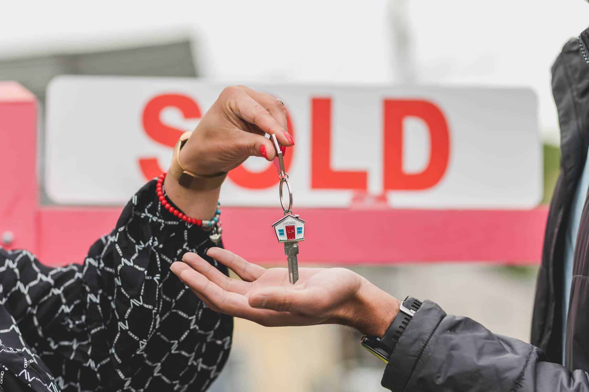 Person handing house keys to another person in front of a sign that says "sold"