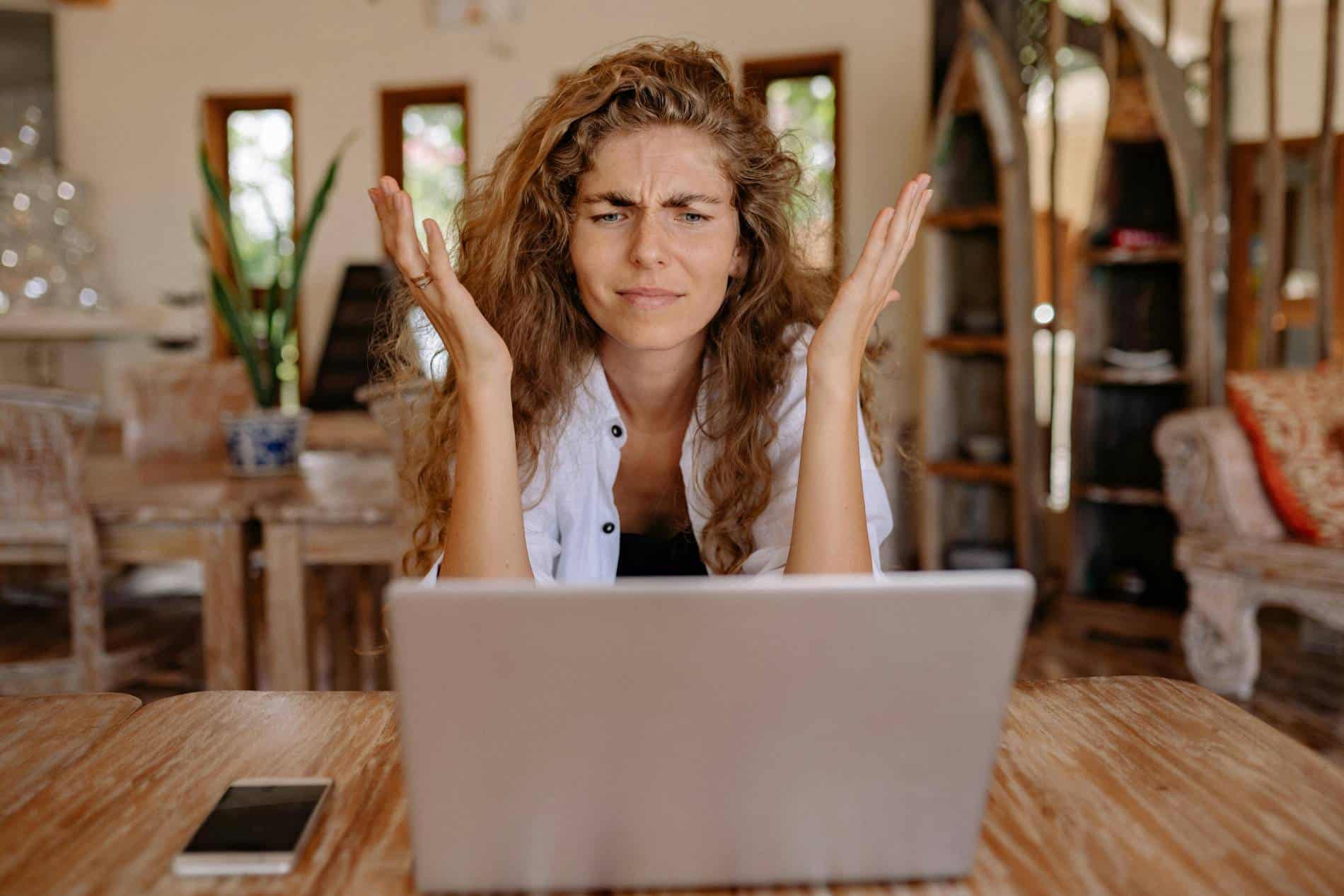 A frustrated looking woman sits in front of a laptop computer, making a face with her hands in the air.