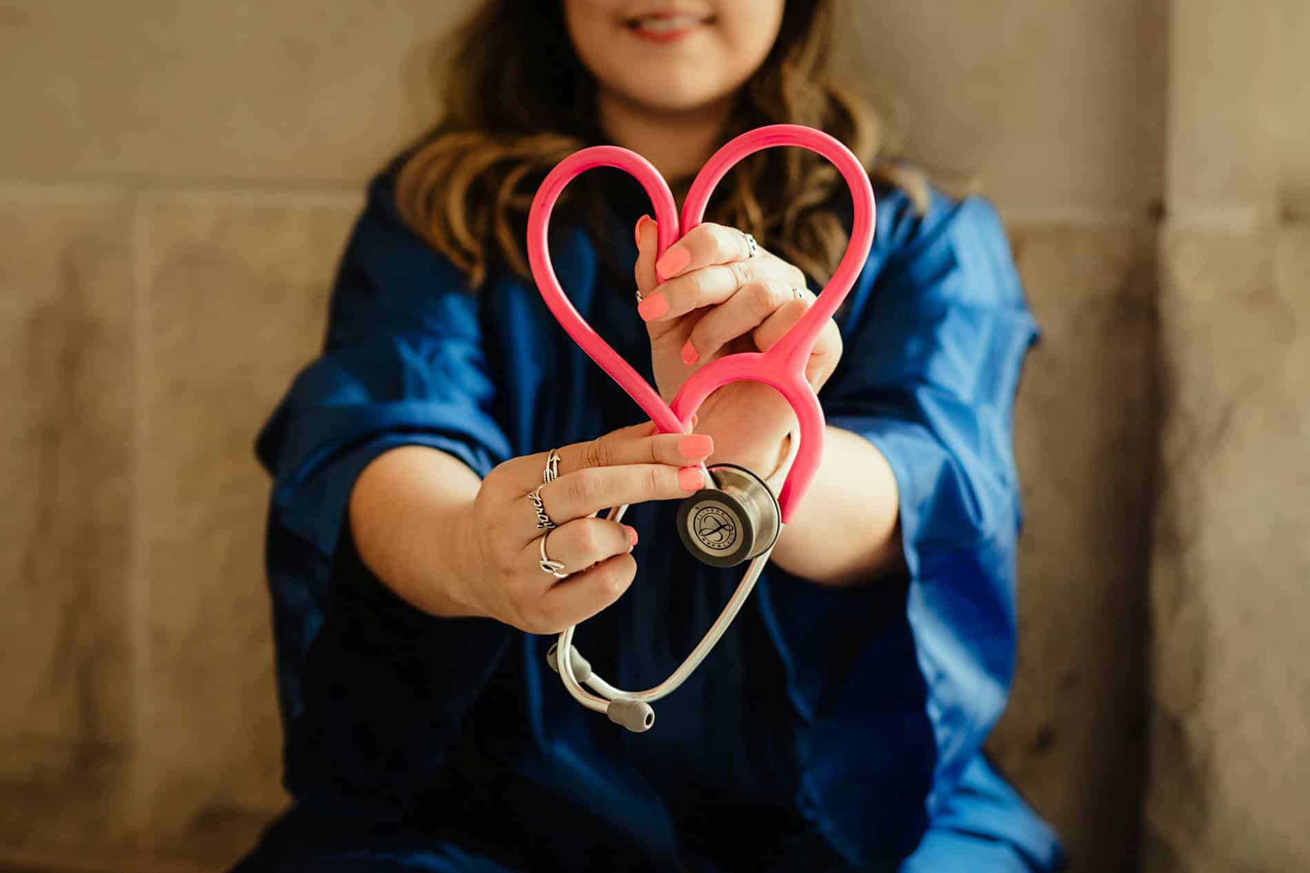 A nursing student in a blue uniform holds a pink stethoscope shaped like a heart
